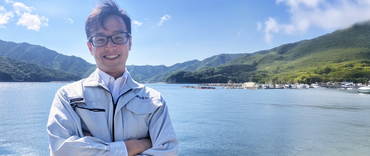 腕を組み立つA.N.さん。後ろに広がるのは米水津湾の海と山