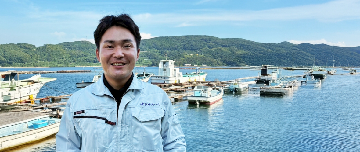 古江湾に浮かぶ漁船らの前で、笑顔で写るF.T.さん