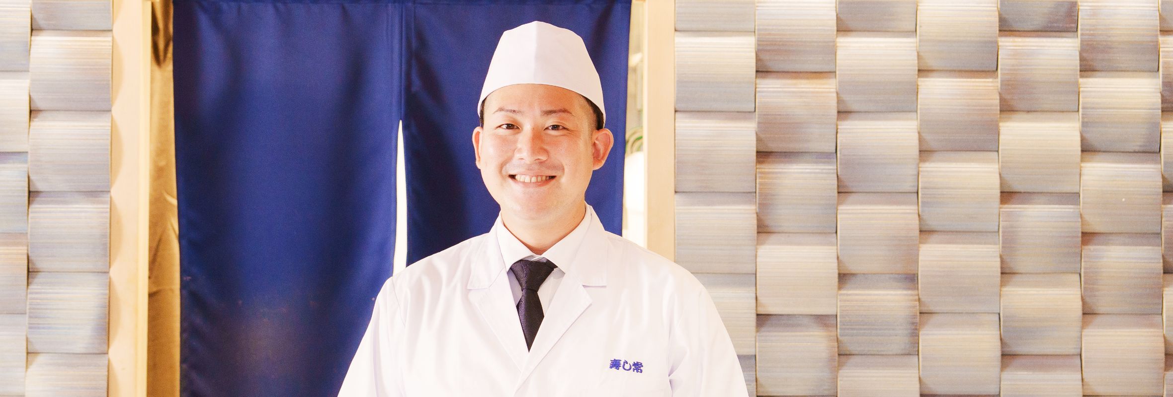 S.Y.さんの正面写真。ネクタイを付けたワイシャツの上から調理白衣を着て、調理帽を身に付けている。背景には半円筒状の白い立体タイルの壁があり、中央には深海色ののれんがかかっている。